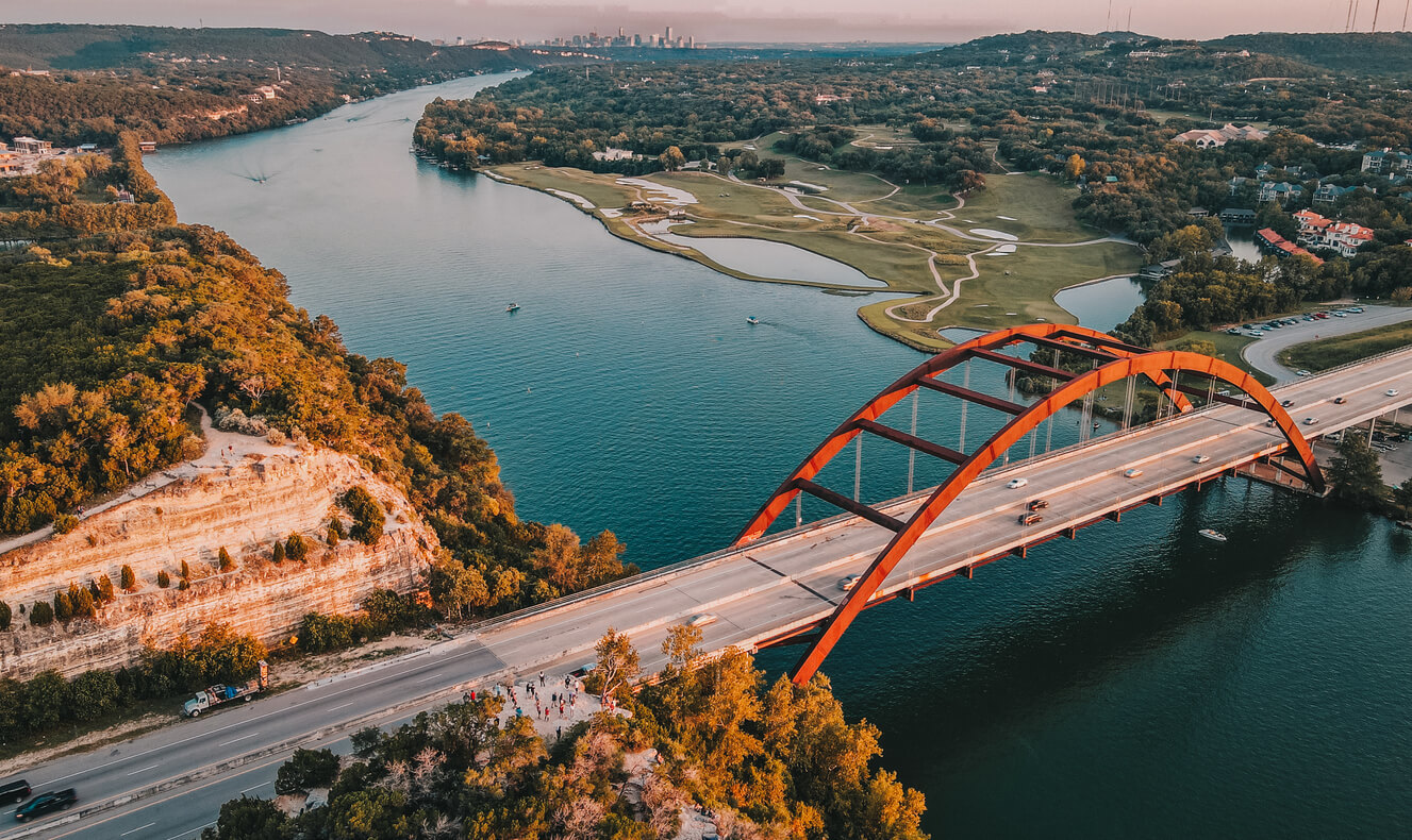 7 Smart Strategies for Buying Austin Waterfront Luxury Homes Aerial view of the Pennybacker Bridge over Lake Austin at sunset with the downtown Austin skyline and Hill Country shoreline in the distance